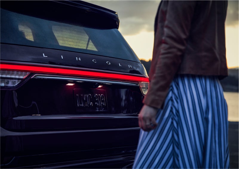 A person is shown near the rear of a 2023 Lincoln Aviator® SUV as the Lincoln Embrace illuminates the rear lights | Bluebonnet Motors Lincoln in New Braunfels TX