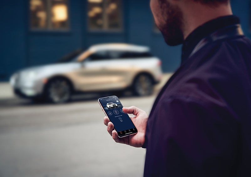 A person is shown interacting with a smartphone to connect to a Lincoln vehicle across the street. | Bluebonnet Motors Lincoln in New Braunfels TX