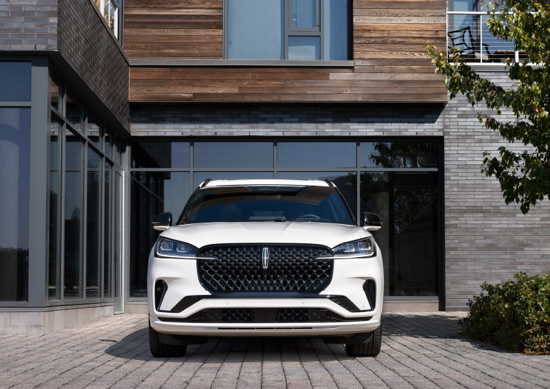 A white 2025 Lincoln Aviator® SUV is shown parked outside a modern building with gray brick and wood accents. | Bluebonnet Motors Lincoln in New Braunfels TX