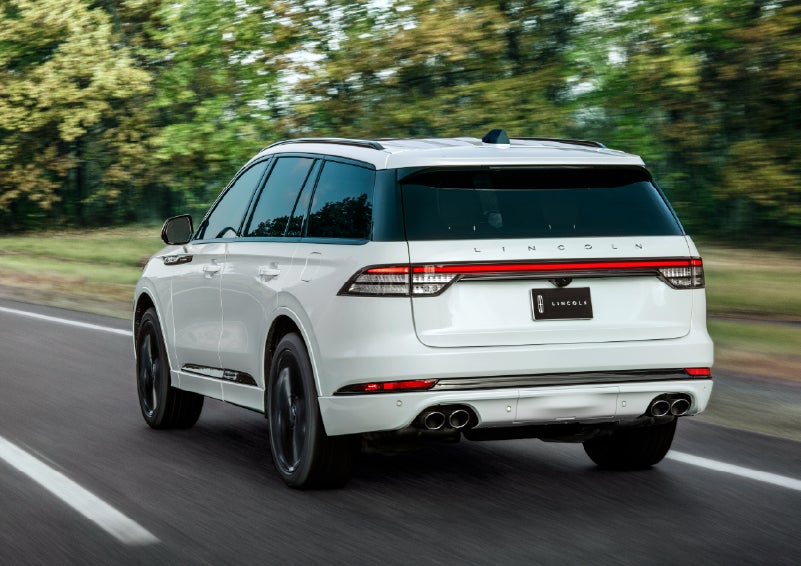 A white 2026 Lincoln Aviator® SUV is being driven away down a road lined with colorful trees. | Bluebonnet Motors Lincoln in New Braunfels TX